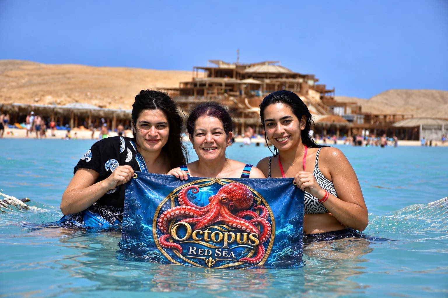 Three women smiling in shallow sea with an Octopus Red Sea branded banner