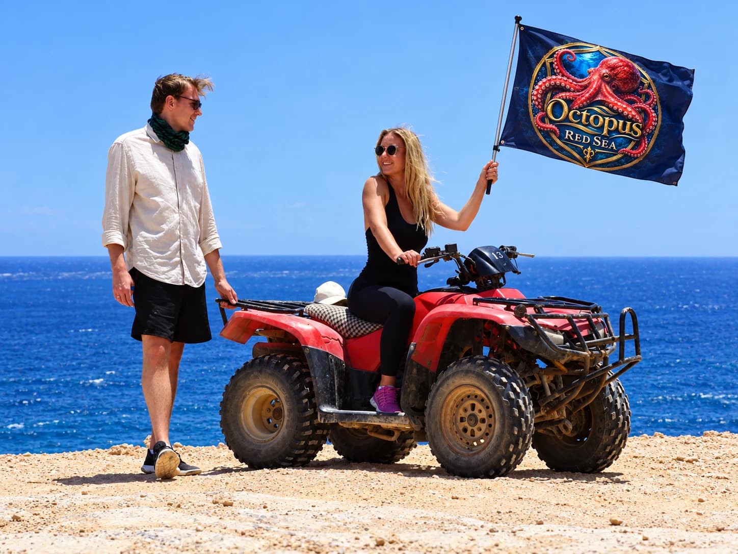 Two travelers posing with a red ATV and an Octopus Red Sea flag by the coast