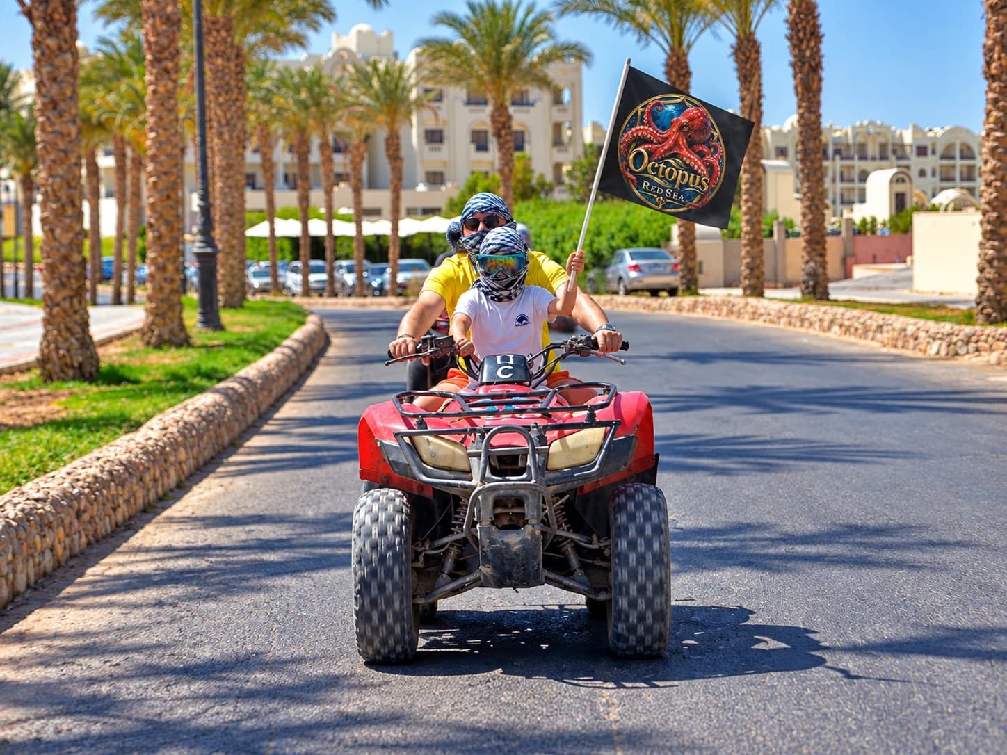 Travelers riding a red quad bike beside palm trees with an Octopus Red Sea flag
